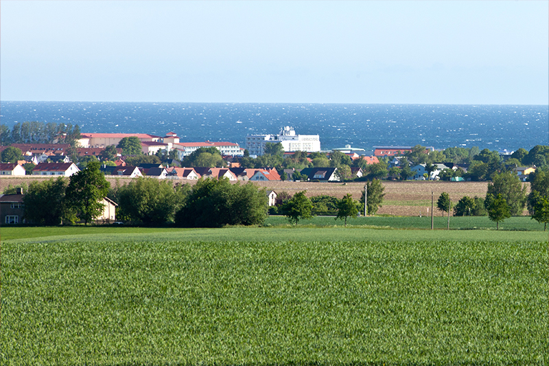 Blick auf eine Küstenstadt mit Feldern und Meer Landschaft mit grünen Feldern im Vordergrund, einer kleinen Stadt in der Mitte und der Ostsee im Hintergrund bei klarem Himmel.