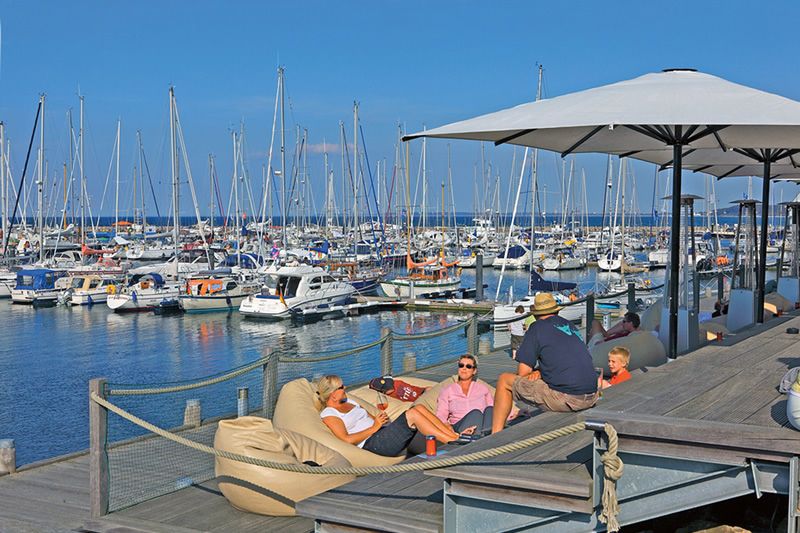 Entspannung am Yachthafen Menschen entspannen sich auf bequemen Sitzmöbeln unter Sonnenschirmen mit Blick auf einen Yachthafen voller Boote.