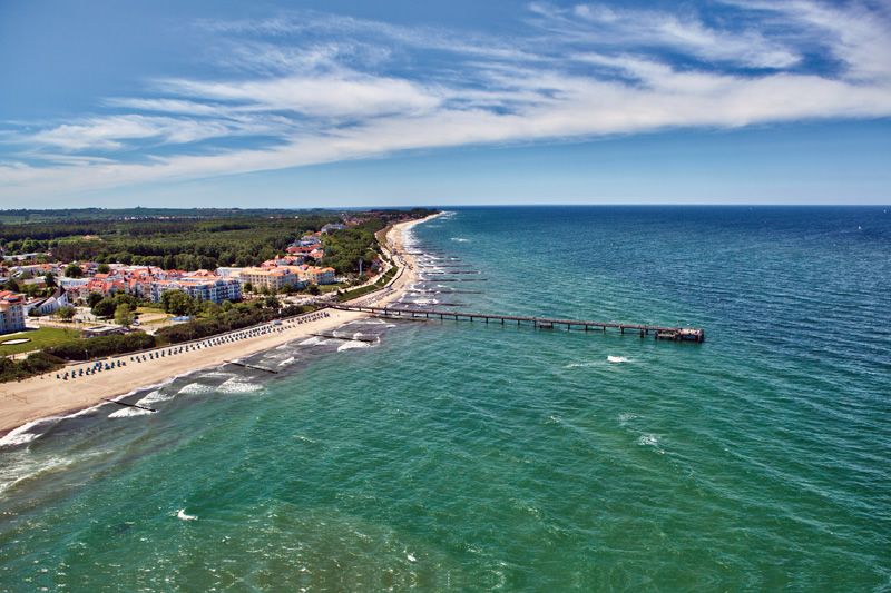 Luftaufnahme der Ostseeküste mit Strand und Pier Luftaufnahme eines Strandes mit Promenade und Pier an der Ostseeküste, mit Gebäuden und Wald im Hintergrund.