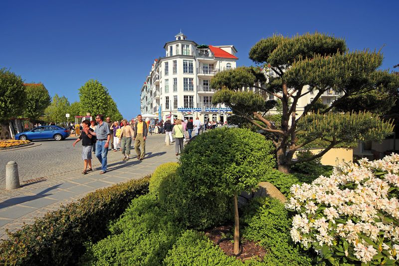 Spaziergang in einer städtischen Umgebung mit moderner Architektur und gepflegter Landschaft. Gruppe von Menschen spaziert auf einer gepflasterten Straße, flankiert von gepflegten, blühenden Pflanzen und einem modernen, mehrstöckigen Gebäude im Hintergrund.