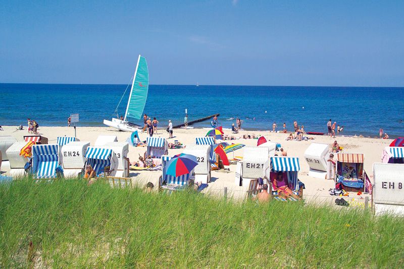 Strandtag mit Strandkörben und Segelboot am Meer Ein sonniger Strand mit Strandkörben, Segelboot und Badegästen, umgeben von blauem Himmel und Meer.