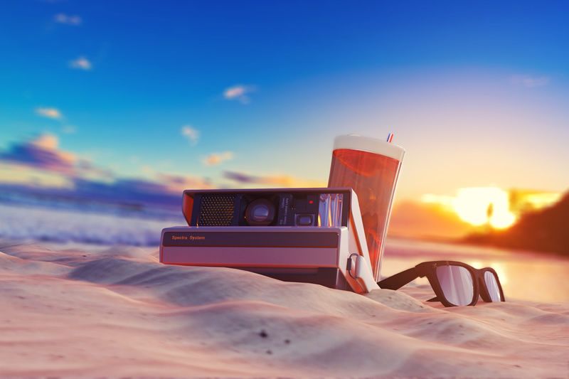 Sommerliche Strandatmosphäre mit Retro-Kamera Polaroidkamera, Getränk und Sonnenbrille im Sand bei Sonnenuntergang am Strand.
