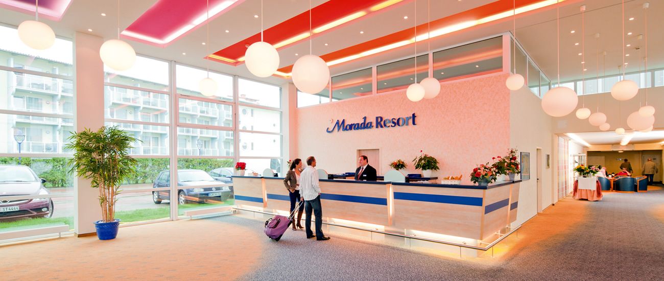 Empfangsbereich eines Resorts mit moderner Einrichtung Helle Hotellobby mit Empfangstheke, an der zwei Gäste mit einem Mitarbeiter sprechen. Große Fenster, moderne Beleuchtung und Pflanzen schaffen eine einladende Atmosphäre.